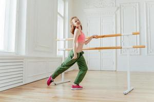 Motivated cheerful red haired female in sports bra and trousers, keeps hand in pocket, wears sneakers, has exercises on ballet barre, dances at studio, being in good mood, finishes dance workout photo