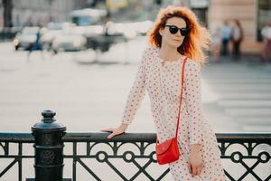 Shot of thoughtful red haired curly woman focused aside, wears long white dress, poses near black hence at street against blurred road with transport, enjoys free time during summer. Lifestyle concept photo