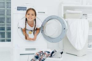 Smiling female child with glad expression, poses in washing machine, dressed in white t shirt, holds detergent, looks gladfully at camera, surrounded with much clothes and linen, does housework photo