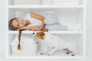 Photo of lovely preschooler lies on white shelf in washing room, has long pigtail, plays with dog, wears casual clothing, smiles gently, spends weekend at home. Children, animals and household concept