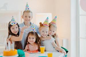 Indoor shot of happy kids celebrate party with falling confetti, wear cone party hats, pose near festive table with cakes, embrace and pose together. Childrens birthday photo