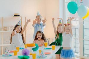 Group of happy children celebrate birthday together, play with confetti, wear party hats, pose near festive table in decorated room with balloons, have overjoyed expressions, enjoy life