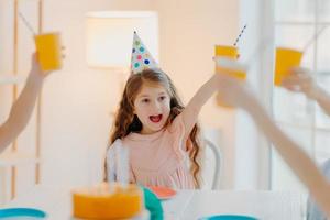 Happy ginger girl wears party hats, clink cups with drink, has fun with friends, celebrate birthday together, pose in decorated room at home. Children, celebration and holiday concept photo