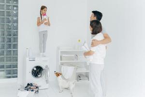 Husband and wife embrace, stand in laundry room, look happily at their single child who stands on washing machine with bottle of washing powder, wash clothes, do family domestic chores at weekend photo