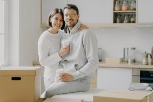 Image of glad family couple hug and stand closely to each other, drink takeout coffee, look with smile at camera, dressed in casual clothes, surrounded with cardboard boxes, spend free time in kitchen photo