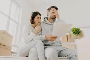 Dejected husband and wife manage finances, receive bill, face financial problem, have gloomy expressions, sit together in empty room, big window behind, cardboard boxes with personal stuff near photo