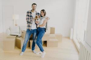 Young couple in love stand closely to each other, embrace and holds favourite dog, pose in empty room, surrounded with cardboard boxes, make family portrait in new apartment. Moving Day concept photo