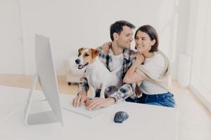 Affectionate couple in love embrace and going to kiss, watch romantic movie at modern computer, pedigree dog focused in monitor with interest. Lovely family spend free time in domestic atmosphere photo