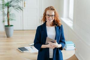 Horizontal shot of proud CEO checks email on digital touchpad, looks at camera with satisfied expression, wears spectacles, stands in modern office, enjoys online communication on modern device photo