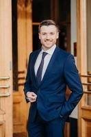 Vertical shot of handsome unshaven male employee has cheerful expression, dressed in elegant suit, stands near entrance of his company, being in good mood, starts new working day, has success photo