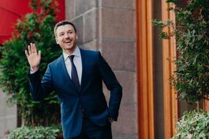 Greeting concept. Cheerful successful young businessman in formal clothes waves with hands as sees colleague into distance, has happy expression, stands outdoor near restaurant. Hey, I am here photo