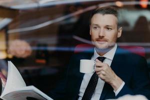 View from window of handsome successful young male boss, holds cup with hot aromatic beverage, looks through menu in luxury restaurant, dressed in formal expensive suit, thinks about something photo