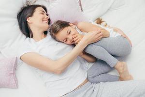 Delighted female child giggles joyfully as plays with her mother in comfortable bed, have positive smiles on faces, wear pyjamas, have good mood after awakening. People, leisure and sleeping concept photo