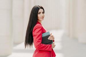 Serious brunette businesswoman with makeup, manicure, wears red jacket, carries diary and notepad, stands in profile outdoor, goes for work. Elegant businesslady on street. Stylish director outside photo