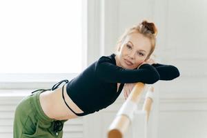 Restful fatigue graceful female dancer practices in dance studio, looks at camera with tired expression, leans at horizontal bars for performing exercises, wears black top and sweats, takes break photo