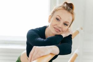 Close up portrait of ginger attractive female dancer with combed hair, makeup, wears black top, leans on barre, has gentle smile, looks straightly at camera. People, choreography, leisure concept photo