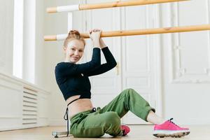 Female gymnast sits on floor, dressed in sport outware, stretches herself near barre, holds hand on handrail, looks with satisfied expression at camera, shows fit body. Dancing, flexibilty, aerobics photo