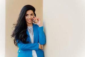 Satisfied brunette business lady calls on smartphone device, books tickets for trip abroad, makes positive conversation, dressed in formal elegant clothes, uses technology, poses near white wall. photo