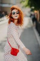 Happy carefree redhead woman laughs positively, spends free time outdoor in city, wears sunglasses, long sleeved dress, carries red bag, has fun, enjoys recreation time. People and lifestyle