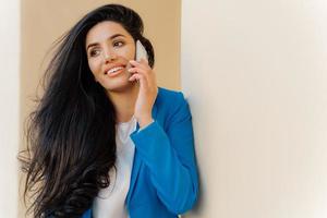 Optimistic female entrepreneur with dark wavy hair, wears makeup, smiles happily, holds modern cell phone near ear has conversation dressed formally looks aside, discusses good news in business sphere photo