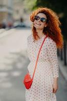 Optimistic foxy woman with glad expression, looks into distance, wears shades, long dress, strolls outdoor on street, enjoys nice summer day, has broad smile. People, lifestyle, rest and lifestyle photo