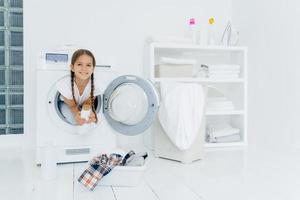 Small girl with appealing appearance, has fun and poses inside of washer, holds detergent, prepares for washing, basin with clothes for putting into washing machine on white floor. Laundry day at home photo