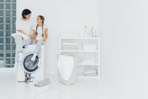 Happy caring woman embraces her little daughter, sit on washing machine, have rest after washing, surrounded with basket and dirty clothes, have pleasant conversation with each other. Housework photo