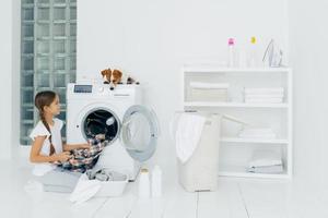 Housekeeping, children and domestic chores concept. Happy kid unloads washing machine, puts clean washed clothes in basin, curious dog looks from above, lies on washer in laundry room at home photo