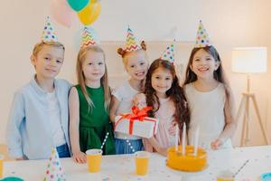 Glad birthday girl stands with presenet, happy friends come to congratulate her, wear party cone hats, stand near festive table with cake, smile joyfully, celebrate festive event photo