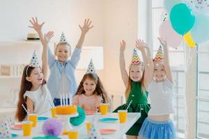 Happy birthday party. Joyful small children have fun together, raise arms and play with confetti, pose at festive table, wear party hats, being on carnival, enjoy holidays