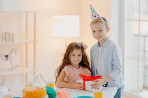 Handsome small boy gives present box to girl, celebrate birthday together, wear festive clothes and party cone hats, pose at table with cake in white spacious room. Holiday concept photo