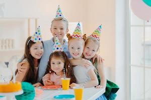 Five friendly little children wear festive cone caps, hug and make photo together, play games and celebrate birthday, have glad expressions, pose at festive table