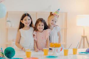 Indoor shot of happy three girls embrace and have fun, smile gladfully, stand near festive table with cake, cups, have happy childhood, being on party together. Childhood and festivity concept photo