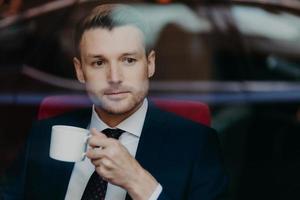 Photo of successful prosperous male entrepreneur with stubble, thinks about something, wears formal clothes, holds cup of coffee, being focused at window, waits for business partner at restaurant