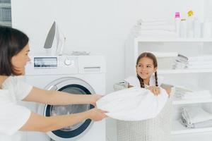 Happy girl with two pigtails poses in basket with dirty linen, has fun in laundry room with mother, helps to do washing. Woman loads washing machine, spend weekend at home, busy with domestic work photo