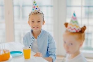 un niño pequeño feliz usa un sombrero de fiesta, se sienta en la mesa cerca de una chica de jengibre, hacen una fiesta juntos, celebran el cumpleaños, posan en una habitación grande y espaciosa con ventana. niños y concepto de vacaciones foto