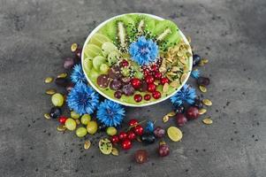 Bowl with fresh kiwi salad, seeds, red current, green grape and blue cornflowers around. Vegeterian dish of ripe fruit. Top view. Blank space photo