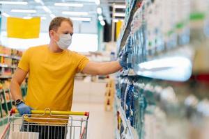 el hombre adulto usa una máscara desechable y guantes de goma como medidas preventivas durante la epidemia, elige agua en la tienda minorista, trata de estar seguro durante el brote de neumonía por coronavirus. autoaislamiento foto