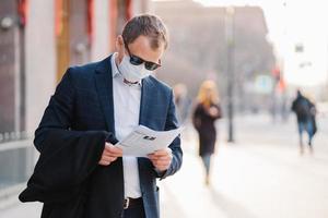 Coronavirus disease. Serious male banker reads newspaper attentively, finds out news about pandemic situation around world, wears protective mask, prevents spreading virus, stands at street. photo
