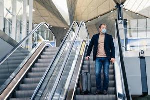 Man wears medical face mask, poses on escalator in airport, arrives from abroad, holds mobile phone and suitcase, protects from pandemic disease. Threat of epidemic in 2020, dangerous traveling photo