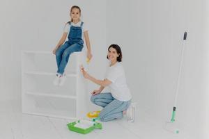 Shot of lovely mother and small hard working daughter pose in empty room, paint furniture in white color, use paintbrushes, happy to finish work, being busy during weekend. People, repair, improvement photo