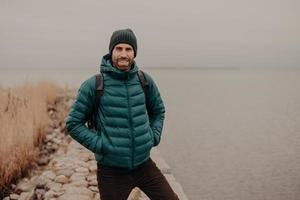 Horizontal shot of attractive unshaven man has cheerful expression, wears hat and jacket, keeps hands in pocket, carries rucksack, has walk during foggy autumn weather, poses near lake outdoor photo