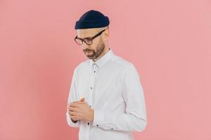 Contemplative unshaven man keeps gaze down, wears white shirt, hat and glasses, being deep in thoughts, tries to make solution, thinks how develop his business, isolated over pink background photo