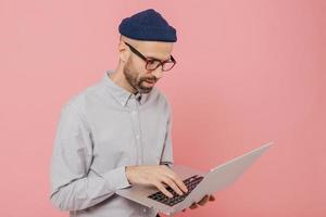 Sideways shot of attractive man stands in front of opened laptop computer, works in internet, wears transparent glasses, white shirt, fashionable hat, models against pink background. Free space photo