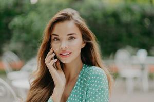 Horizontal shot of pretty young woman wears polkadot blouse, makes phone call, speaks with friend, poses outside against green blurred background, wears lipstick. People and communication concept