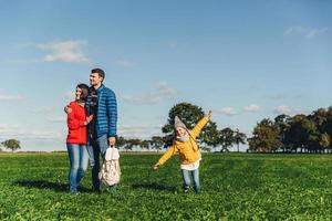 Playful little child runs on green meadow, plays near parents, who embrace each other, look into distance with thoughtful expressions. Family has walk or stroll in countryside, rest after noise city photo