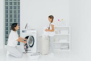 Busy brunette woman loads washing machine with dirty clothes, her little daughter helps, stands near basket and sorts laundry. Happy mother and child in washing room. Household work concept.
