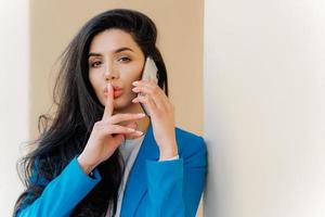 Mysterious brunette woman with makeup, talks with business party, tells secret, makes silent gesture, looks straightly at camera, wears formal apparel. People, communication and conspiracy concept photo