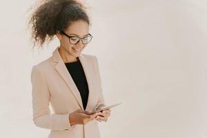 Happy business lady stands with touchpad, reads news online, wears spectacles and elegant clothes, has curly hair combed in pony tail, stands over white wall with blank space, looks for information photo