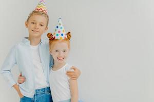 Isolated shot of happy broher and sister cuddle each other, have positive expressions, wear party hats, going to celebrate birthday, stand against white background, copy space aside. Cheerful children photo